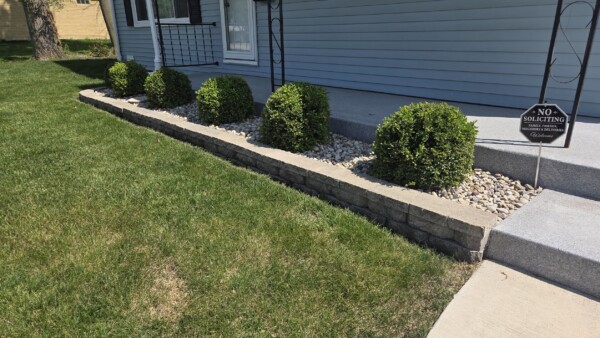 A row of trimmed green bushes lines a stone-bordered garden bed beside a concrete porch, with a No Soliciting sign and a neatly mowed lawn.