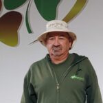 A man wearing a light bucket hat and a green zip-up hoodie stands in front of a wall with a green and brown abstract design.