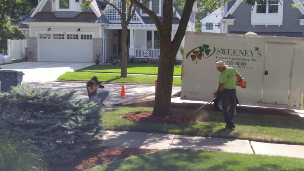 A landscaper in a green shirt uses a leaf blower near a tree, while a photographer kneels nearby taking pictures. A Sweeneys landscaping trailer is parked on the street in a suburban neighborhood.
