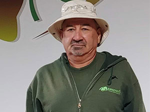 A man wearing a light-colored bucket hat and a green hoodie stands in front of a white wall with a green and brown logo.