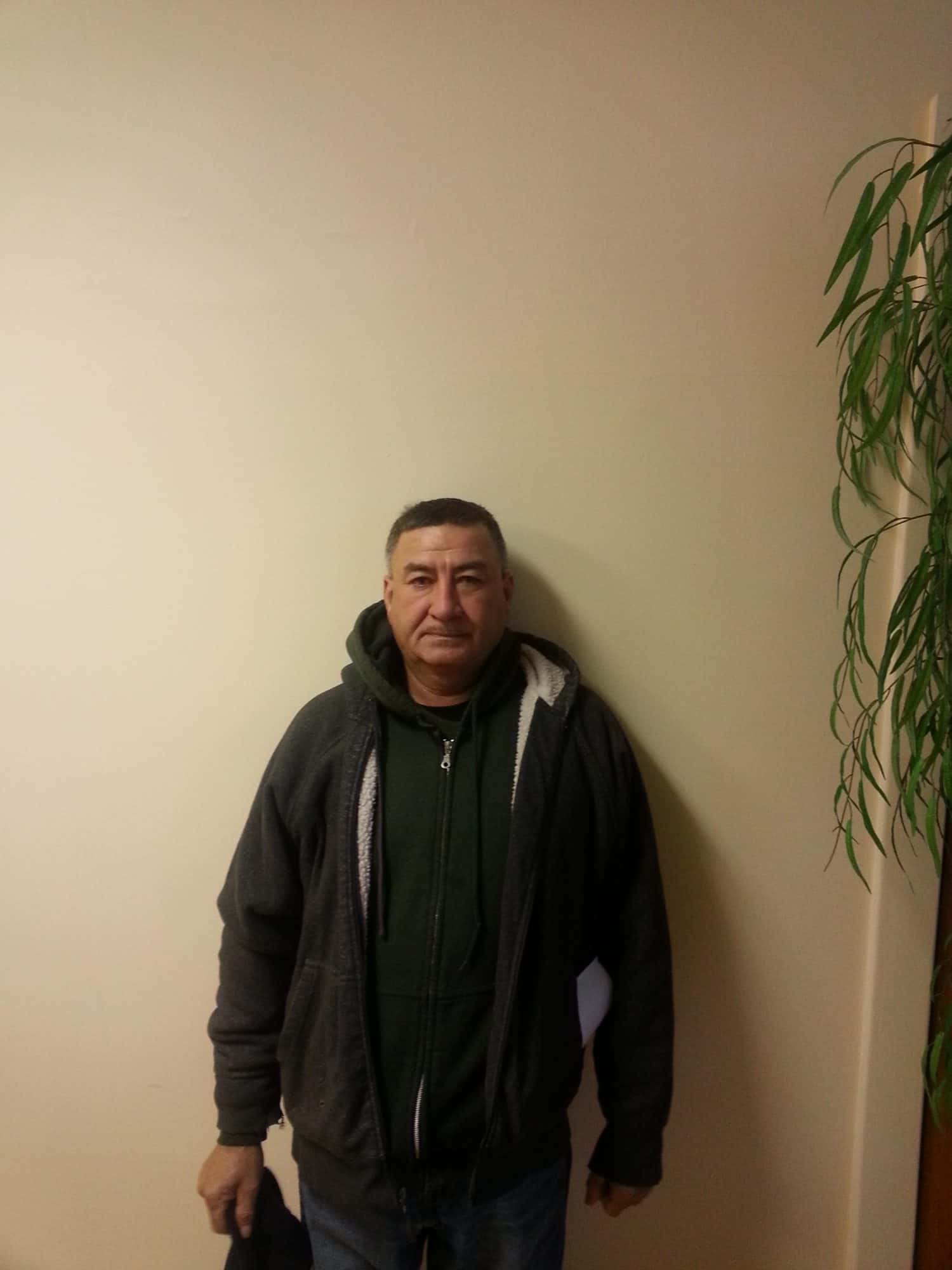 A man with short hair, wearing a dark hoodie over a green zip-up jacket, stands against a plain beige wall. A leafy plant is visible on the right edge of the image.