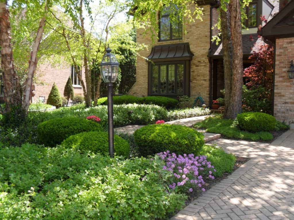 A landscaped front yard with green bushes, blooming flowers, a brick pathway, a lamp post, and a brick house partially visible in the background. Sunny weather with dappled light on the plants.