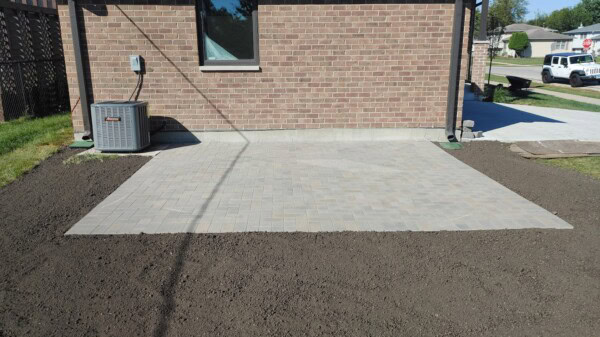 A newly installed light-colored paver patio sits beside a brick house, bordered by freshly tilled soil and a grassy lawn. An air conditioning unit is placed to the left of the patio.