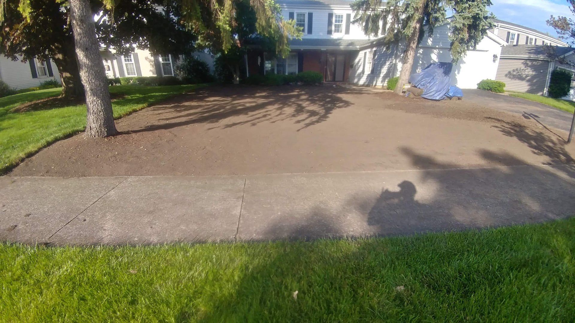 A yard with bare soil in front of a house, bordered by grass and a sidewalk. Trees cast shadows over the area, and there is a blue tarp covering something on the right side.