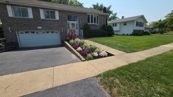 A brick house with a double garage, a driveway, and a flower bed with colorful flowers near the front steps. The lawn is neatly mowed, and a sidewalk runs along the street.