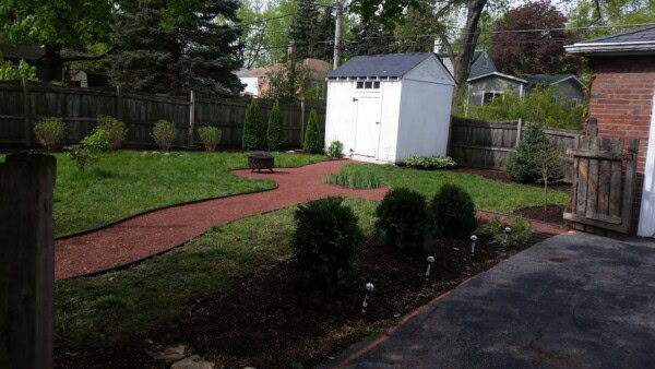 A backyard garden with a winding red gravel path, small bushes, lawn, and a white shed surrounded by a wooden fence. Some solar lights line the path near the driveway.