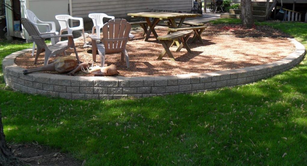 A backyard patio with stone edging features picnic tables and plastic chairs on gravel, surrounded by green grass and shaded by nearby trees.