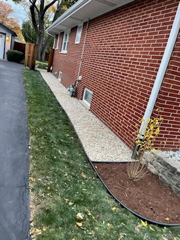A brick house with a gravel pathway runs alongside it, bordered by a narrow strip of grass and a small mulched area with a young shrub.