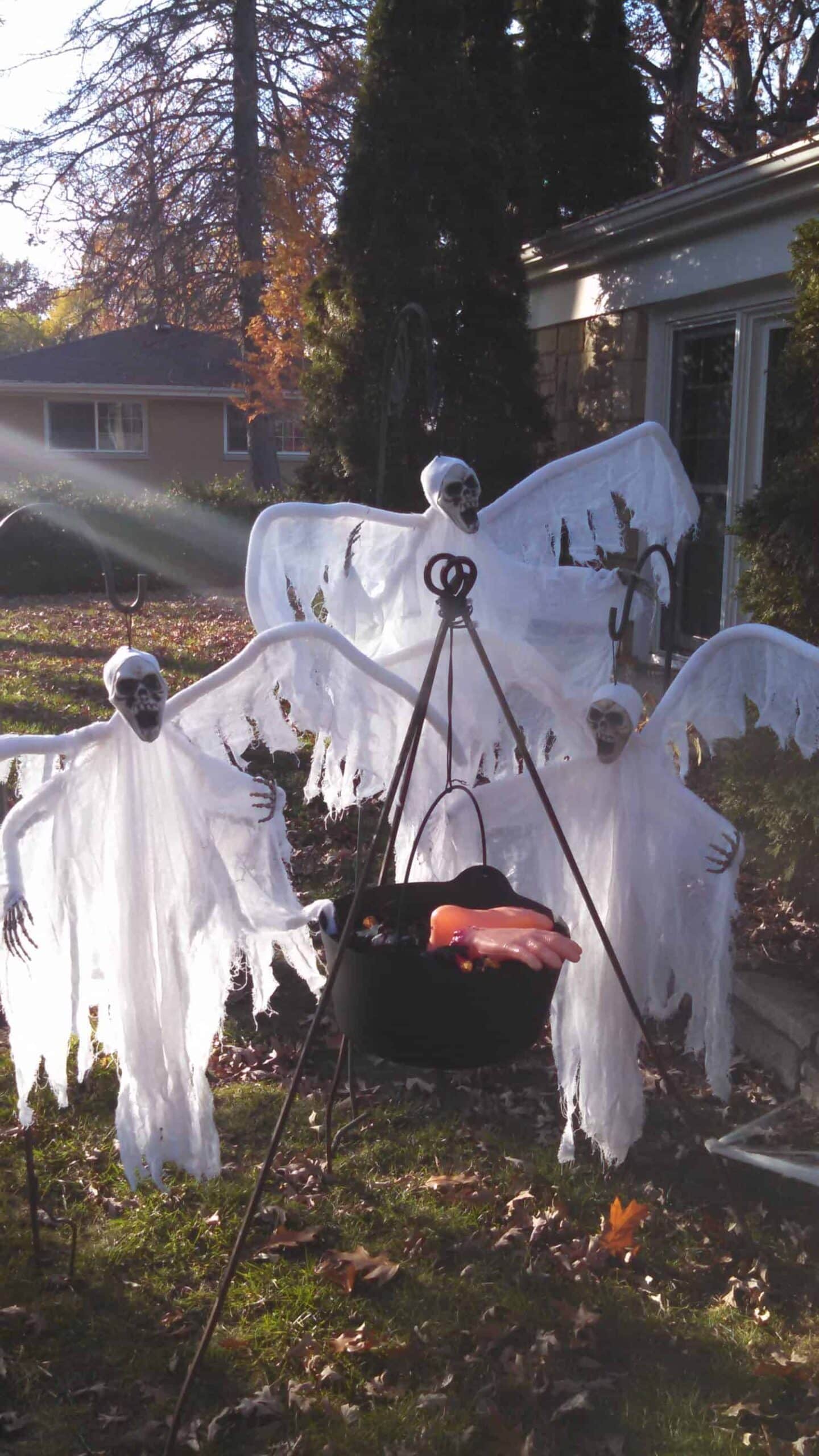 Three ghost decorations stand around a black cauldron with a fake severed hand inside, set up outdoors on a lawn with autumn leaves and trees in the background.