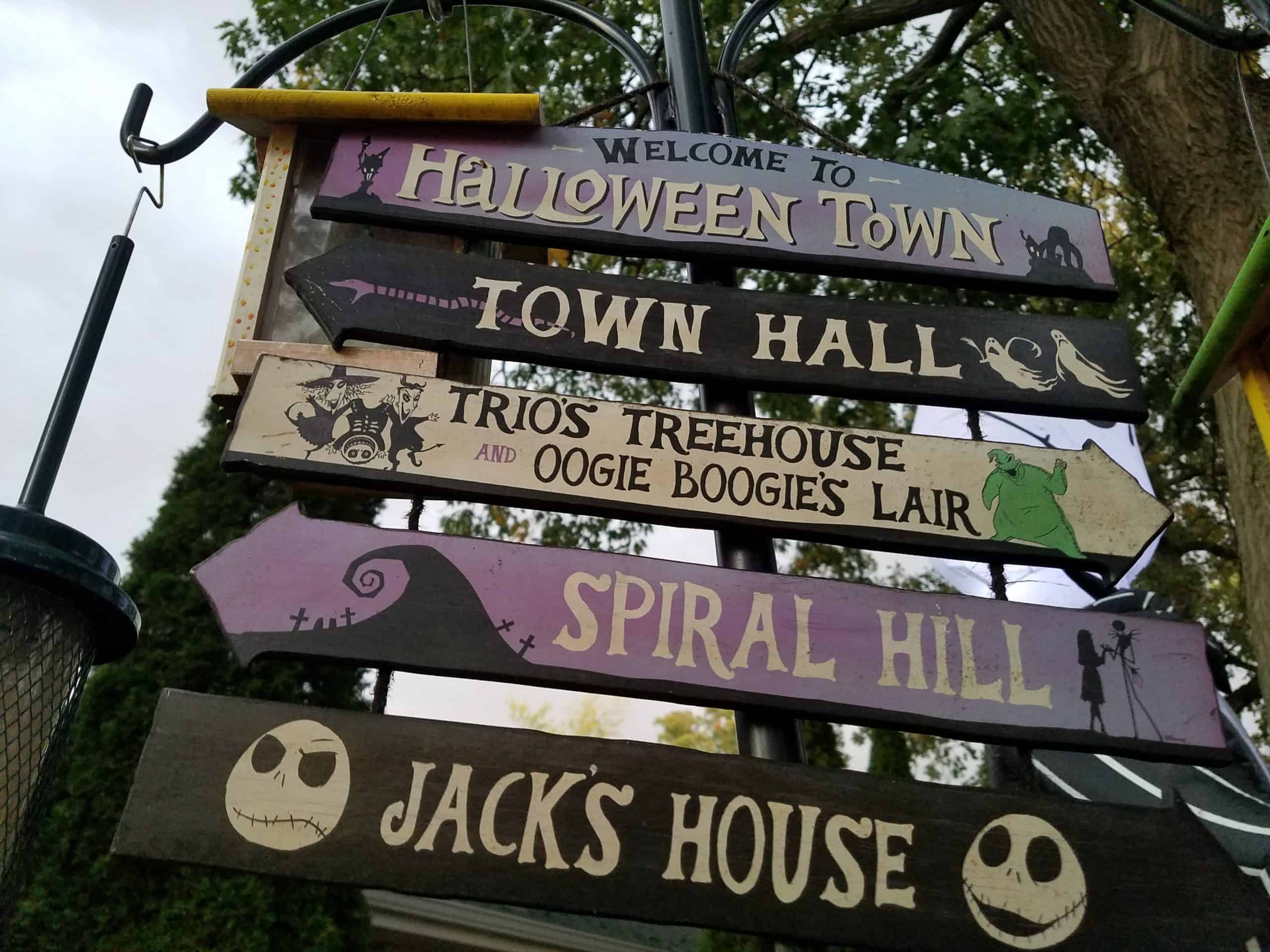Five whimsical wooden signs point in different directions. They read: Halloween Town, Town Hall, Trios Treehouse and Oogie Boogies Lair, Spiral Hill, and Jacks House, with themed illustrations.
