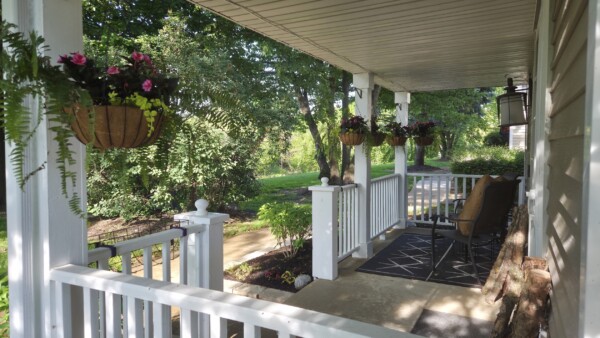 A sunny porch with white railings, hanging flower baskets, a black rug, and cushioned chairs overlooks a lush green yard and garden path.