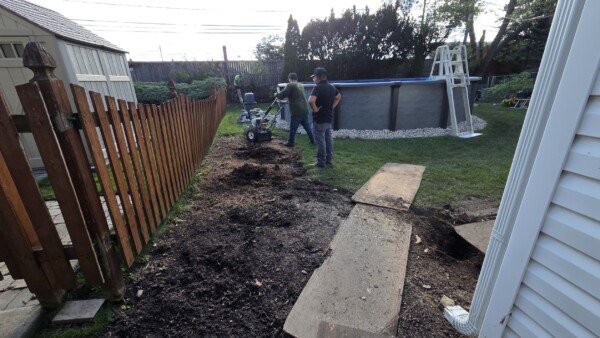 Two people stand in a backyard near an above-ground pool, next to a wooden fence and boards on the ground, with gardening equipment and dug-up soil visible.