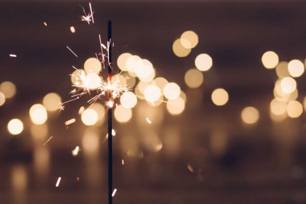 A lit sparkler emitting bright sparks, with warm, blurred lights glowing softly in the background.