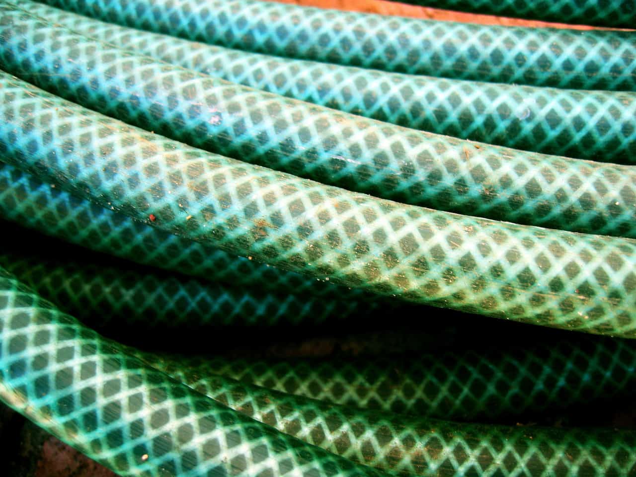Close-up of several green garden hoses with a crisscross diamond pattern, coiled and overlapping each other. The hoses appear slightly worn and are arranged diagonally across the image.