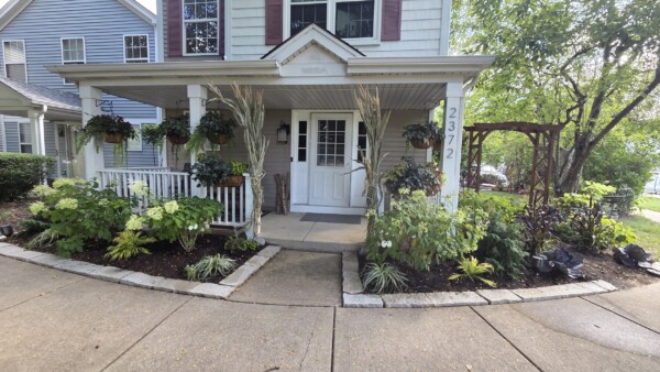 A house with a white porch, potted plants, and hanging baskets, surrounded by landscaped gardens and a curved driveway. The address 23772 is visible on a porch post. Trees and greenery are in the background.