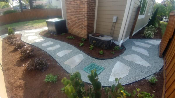 Curved stone pathway with large stepping stones set in gravel runs alongside a house, bordered by mulch and landscaped plants, with two air conditioning units near the brick wall.