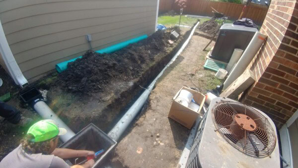 A person installs drainage pipes along the side of a house, with soil mounds and boxes nearby. An air conditioning unit sits next to the work area.