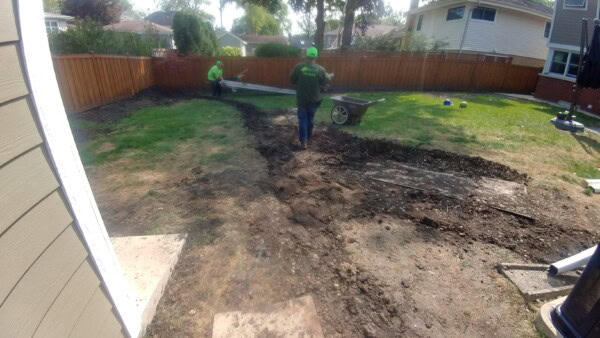 Two workers in green shirts landscape a backyard, moving dirt and using tools. A wheelbarrow sits nearby, and the yard shows patches of grass, dirt, and dug-out areas. Houses and a wooden fence are in the background.