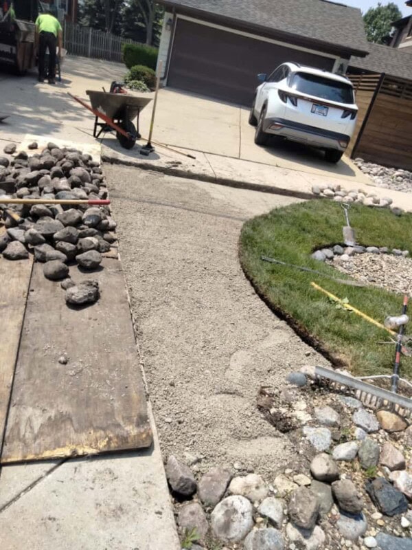 Fresh concrete is being poured for a pathway next to a driveway. There are rocks, a wheelbarrow, and construction tools nearby. A white car is parked in the driveway.