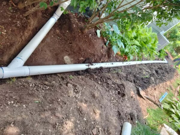 Large PVC pipes are laid in a trench in a garden area, surrounded by soil, plants, and some gardening tools.