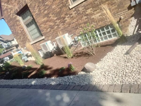 A mulched garden bed with small plants, ornamental grasses, rocks, and two air conditioning units next to a brick building. A stone border separates the garden from a gravel area and sidewalk.