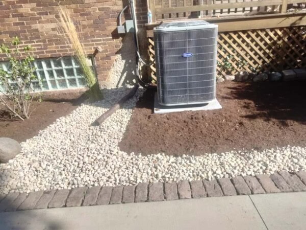 An air conditioning unit sits on a concrete pad beside a house, surrounded by fresh soil, decorative rocks, and landscaping under a wooden deck railing.