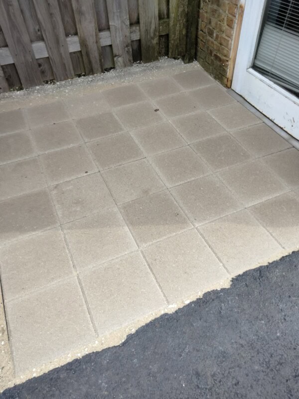 A newly installed square-tiled patio area next to a wooden fence and a glass door, with sand visible around the tiles and adjacent black asphalt.