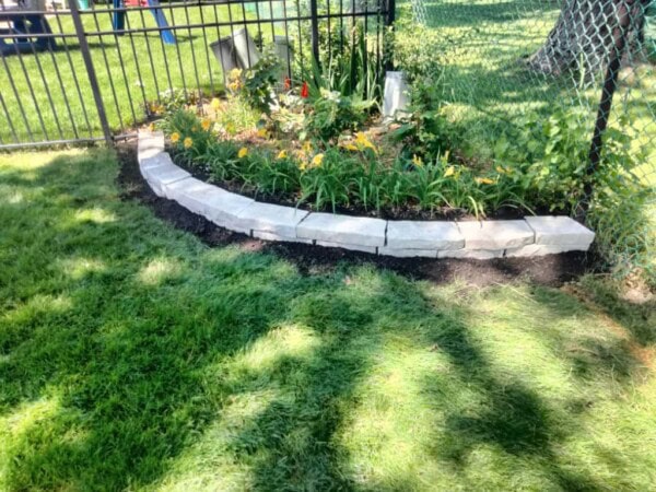 A curved flowerbed with yellow flowers and greenery is bordered by light stone bricks, next to a black metal fence on a sunny, grassy lawn.