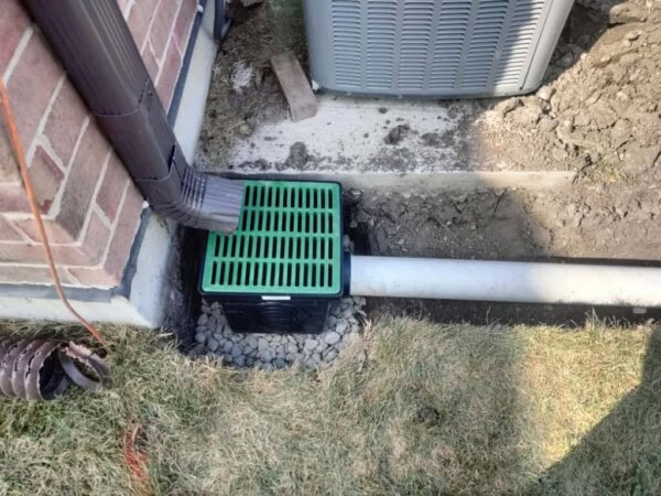 A green drain grate connects to a downspout beside a brick house, directing water into a white underground pipe. Gravel surrounds the grate, and some grass and soil are visible nearby.