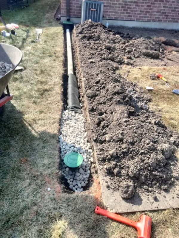 A partially covered French drain beside a house shows a black pipe, gravel, and soil, with tools scattered nearby, including a red mallet and a wheelbarrow.