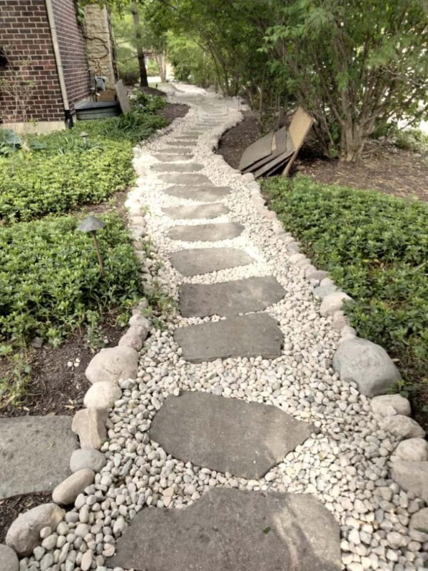 A curving garden path made of large flat stones set in white gravel, bordered by round rocks, winds through greenery and bushes beside a brick house.