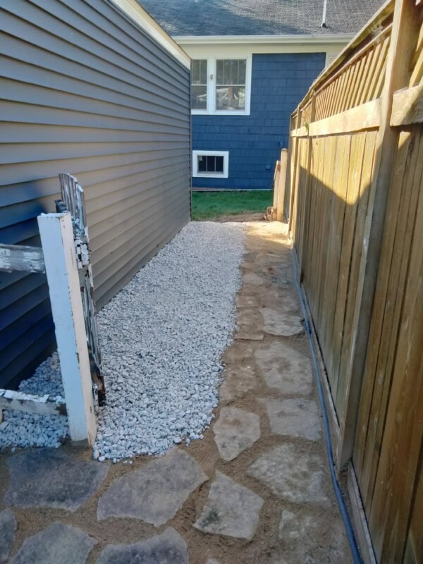 A narrow backyard pathway with stone pavers on the right and a section of white gravel on the left, bordered by a wooden fence and a blue house.