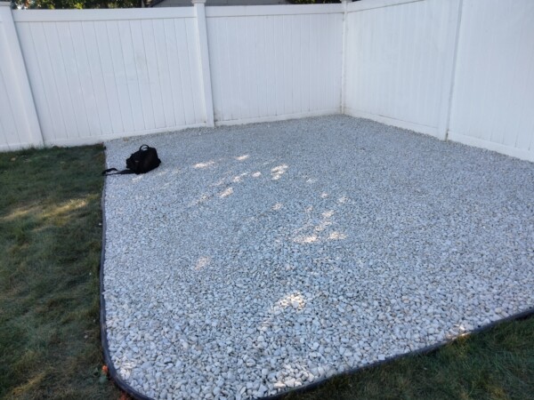 A fenced backyard corner with white gravel covering the ground, bordered by grass on one side. A black bag sits on the gravel near the grass edge.