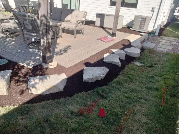 Large stones border a mulched garden bed next to a patio with outdoor furniture. A small red flag and orange markings are on the grass, and sunlight casts shadows across the area.