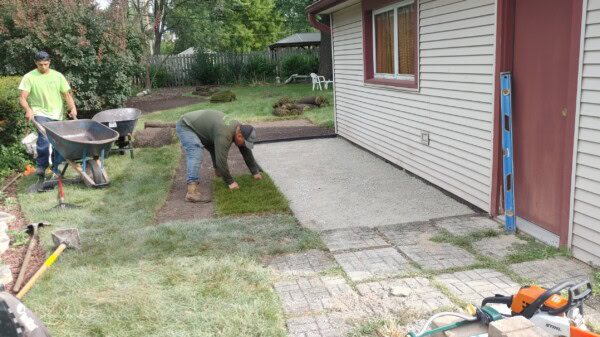 Two workers lay sod next to a house patio. One spreads grass while the other stands near wheelbarrows. Tools and construction materials are visible around the area.