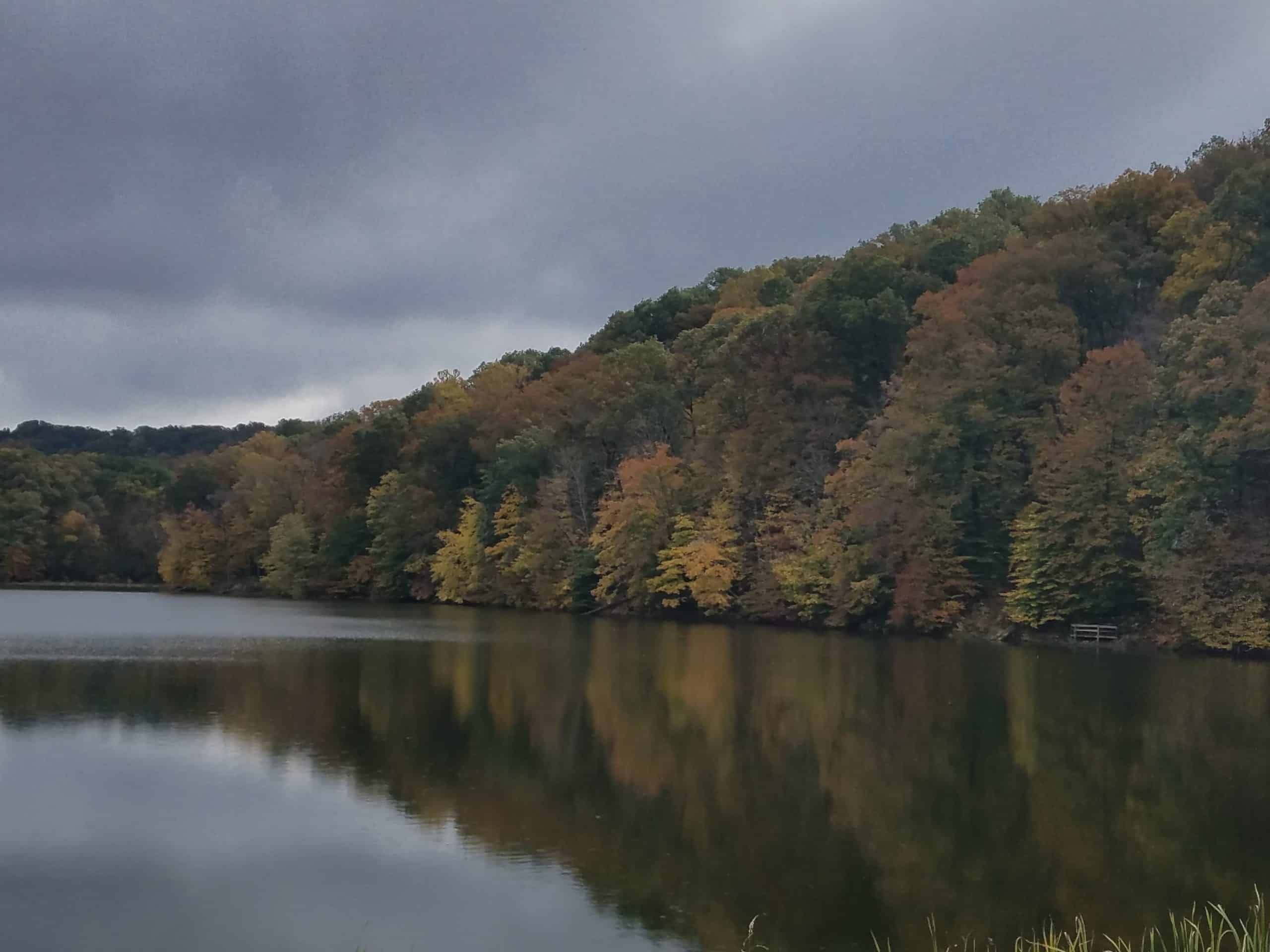 Cloudy sky over a calm lake reflecting a tree-covered hillside with autumn foliage in shades of green, yellow, and orange.