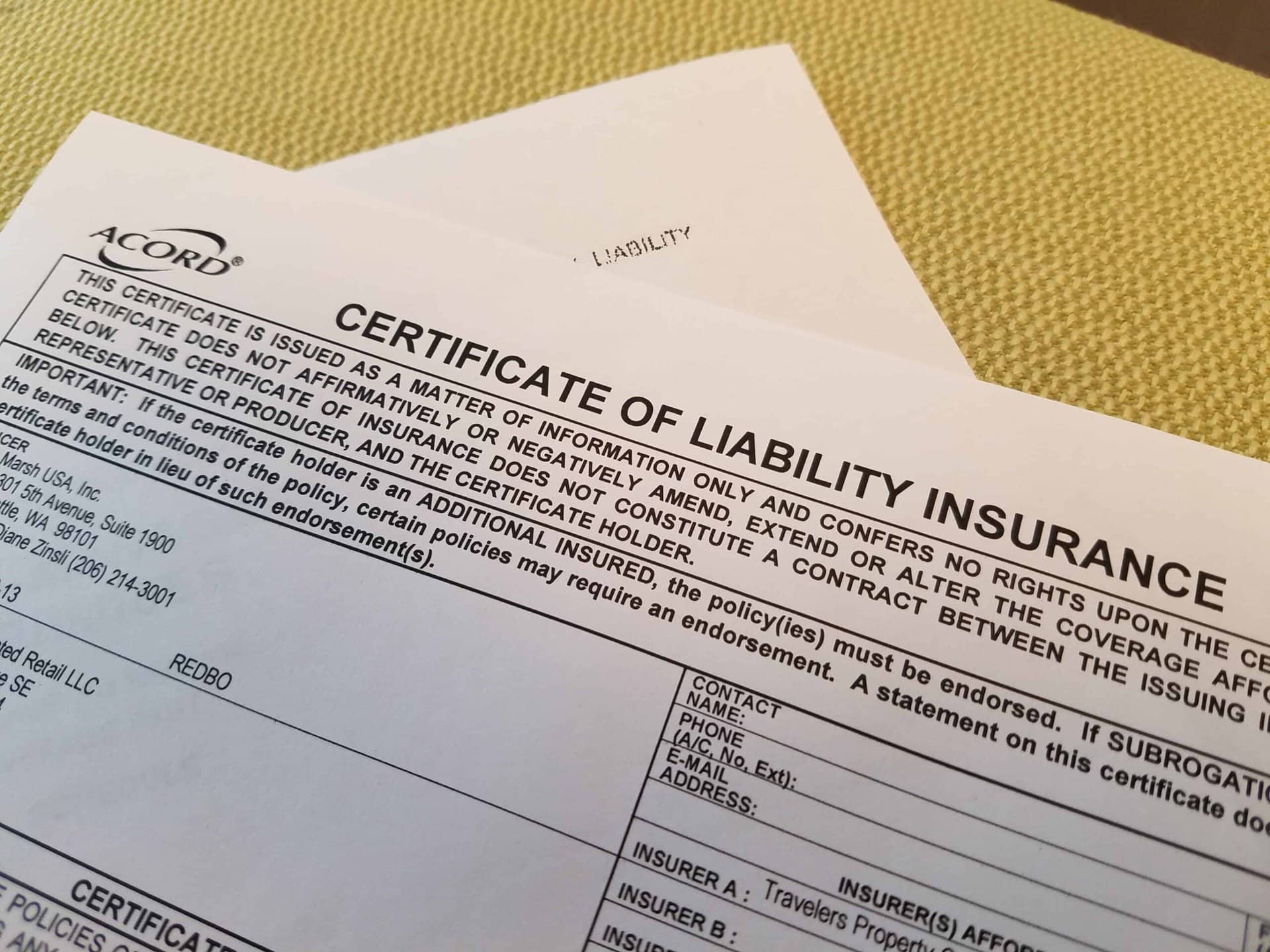 A close-up of a Certificate of Liability Insurance form on a table, with part of another document partially visible underneath. The form contains text and fields to be filled out.