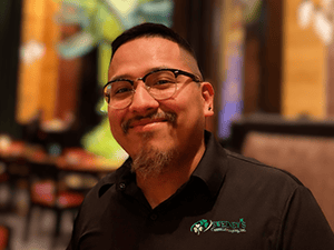 A man with glasses and a goatee smiles at the camera in a restaurant. He wears a black collared shirt with a green and white embroidered logo on the chest. The background is softly blurred.