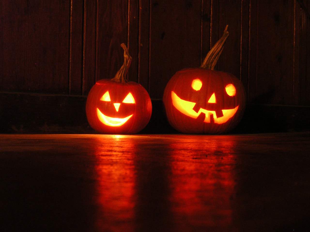 Two glowing jack-o-lanterns with carved faces sit on a wooden floor, casting reflections and an orange glow against a dark wooden background.