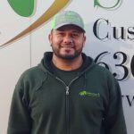 A man wearing a green zip-up hoodie and green cap with a logo, standing in front of a company sign with green and gold graphics and partial text visible.