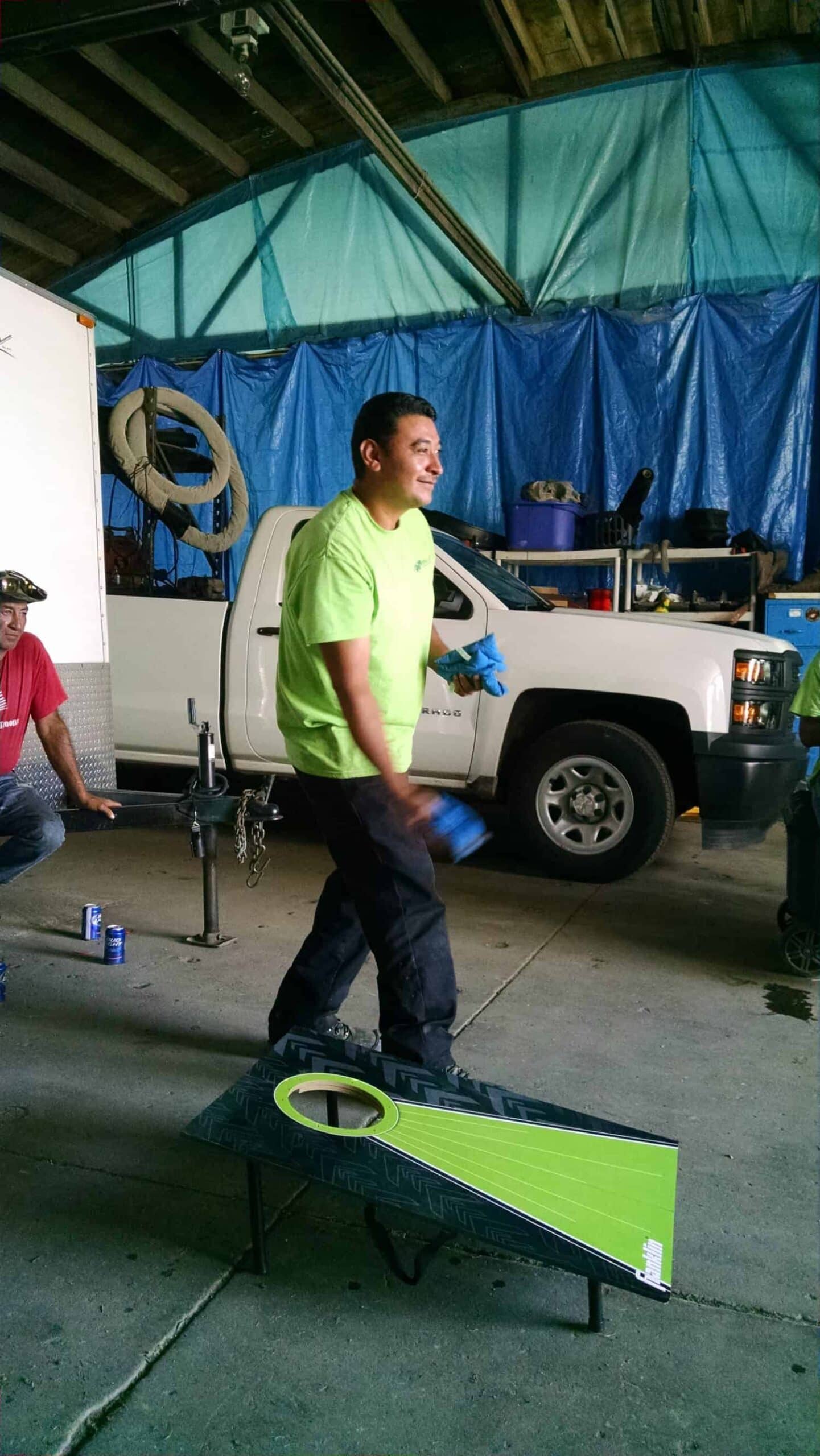 A man in a bright green shirt and gloves smiles while playing cornhole indoors, with a white truck and blue tarps in the background. Two other people are sitting nearby, watching him.