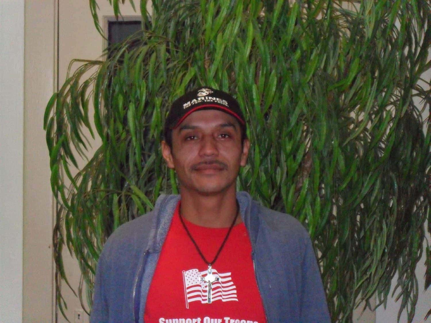 A man wearing a red Support Our Troops shirt, blue jacket, and black Marines cap stands in front of a tall green plant, facing the camera.