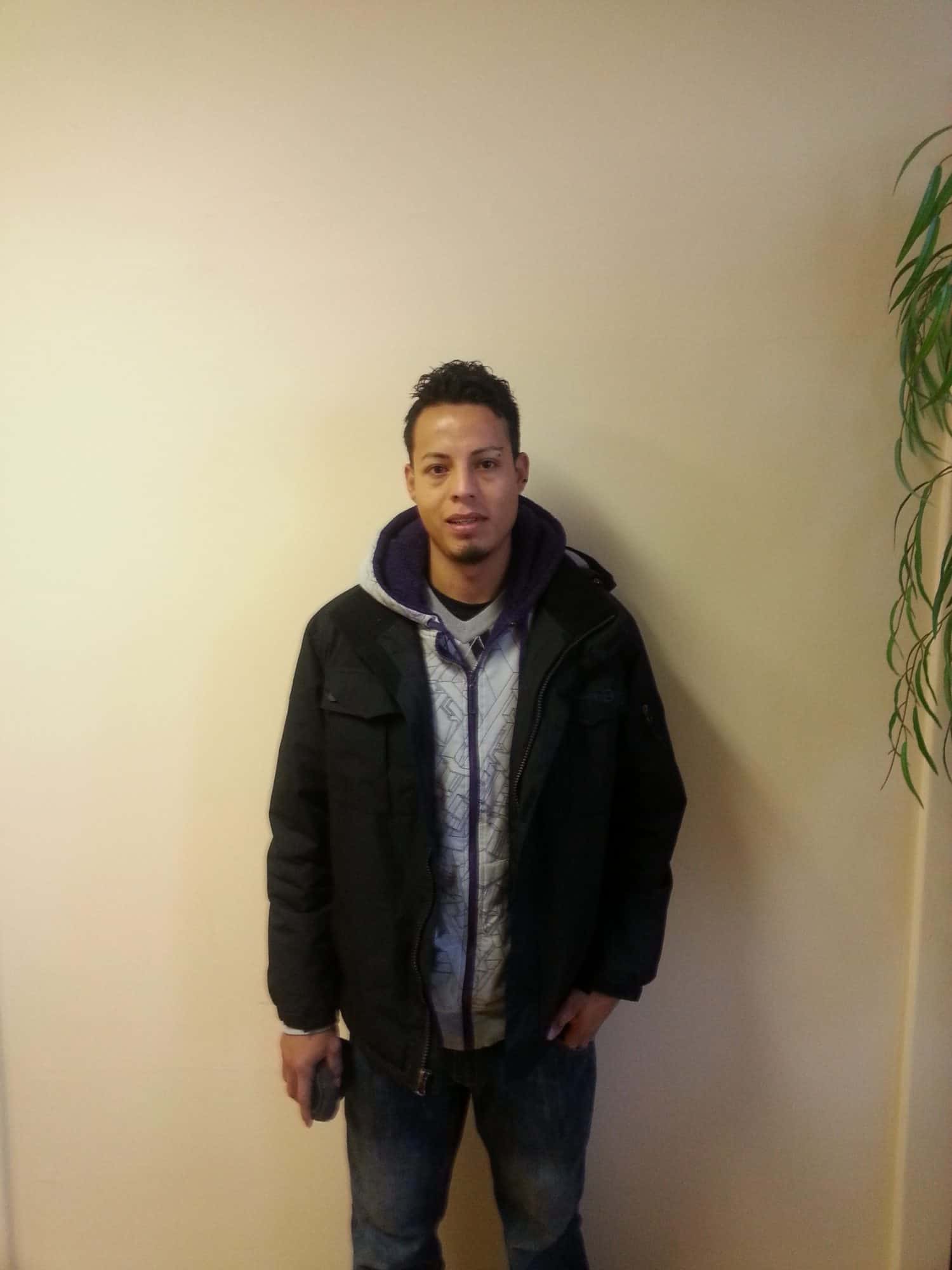 A man stands against a beige wall, wearing a black jacket over a gray hoodie and jeans. He holds a phone in his right hand. A leafy plant is partially visible on the right side of the image.