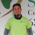 A man wearing a yellow-green shirt and gray sleeves stands smiling in front of a white wall with green and gold graphics and partial text.