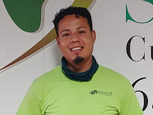A man with short dark hair and a goatee smiles at the camera, wearing a light green shirt and a dark neck gaiter. A patterned wall with text is in the background.