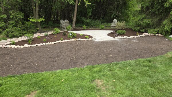 A landscaped garden area with a stone path, mulched flower beds, two wooden chairs in the background, and a border of rocks, surrounded by green grass and trees.