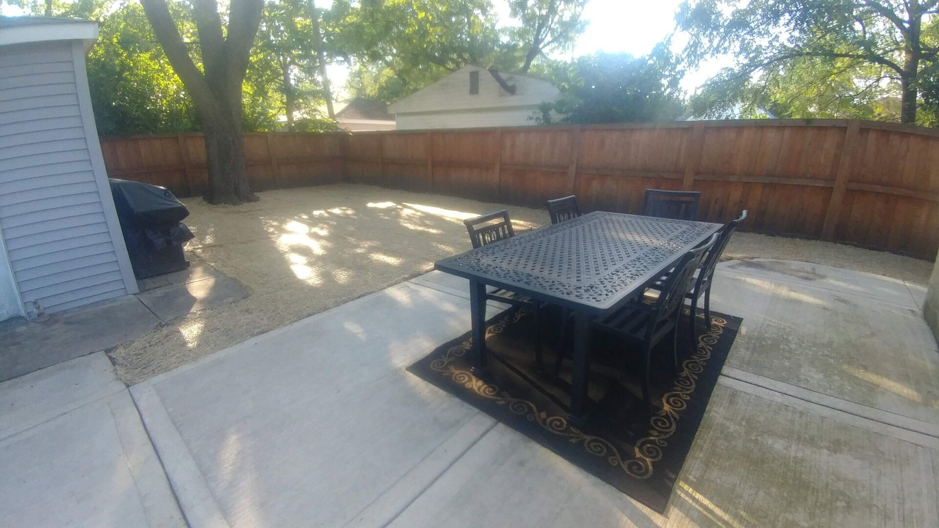 Outdoor patio with a black metal dining table and six chairs on a rug, set on a concrete slab. The yard is fenced with trees, and there is a grill to the left near the fence.