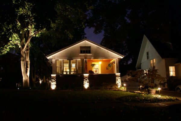 A cozy house is warmly lit at night, with lights illuminating the porch, walls, and nearby trees. A large tree stands on the left, while another house is partly visible on the right.