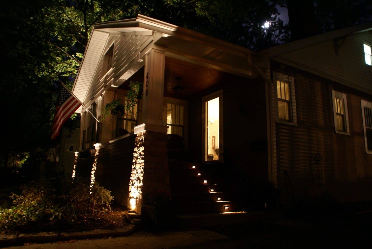 A house at night with lights illuminating the front steps and porch, an American flag hanging, and light visible through the windows. Trees and a faint moon are seen in the background.