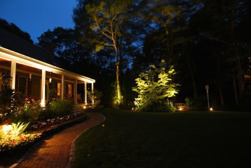 A house at night with a lit porch, brick walkway, garden beds, and trees illuminated by outdoor landscape lighting in the yard.